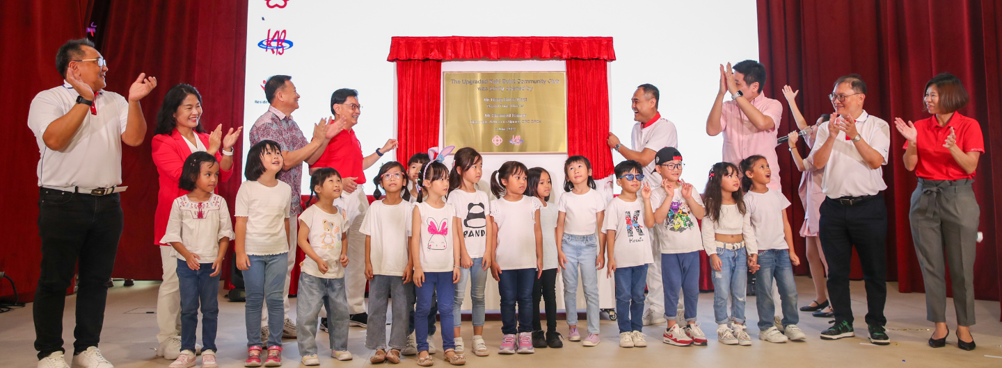 Group of children and adults standing before a plaque and red curtain, clapping.